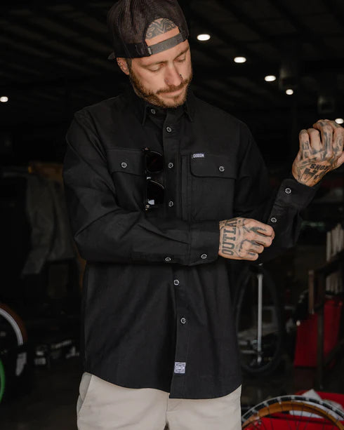 MEN'S LONG SLEEVE WORKSHIRT (Black) | DIXXON worn by a tattooed man in an industrial setting, showcasing performance and style.